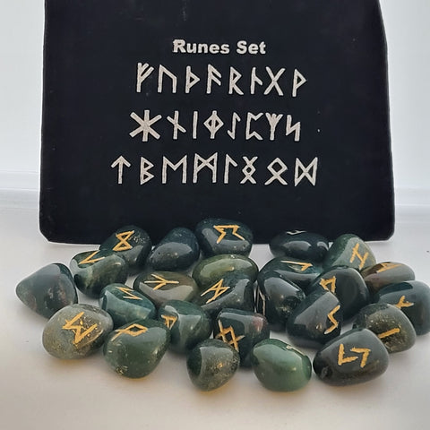 Bloodstone rune stones with golden symbols scattered in front of a black pouch featuring runic designs, showcasing their smooth surfaces and deep green with red speckled coloring.