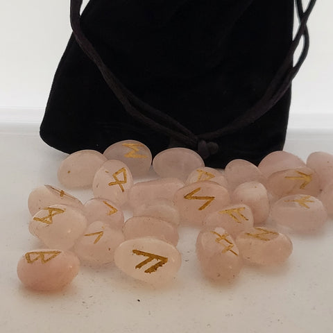 Rose quartz rune stones with golden symbols scattered in front of a black pouch featuring runic designs, showcasing their smooth, polished surfaces and soft pink color.