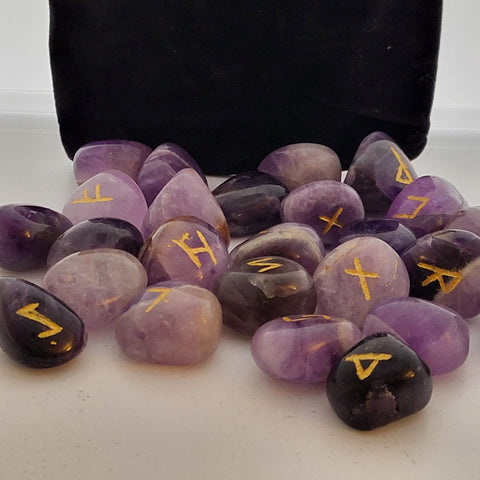 Amethyst rune stones with golden symbols scattered in front of a black pouch, showcasing their smooth, polished surfaces and mystical markings.