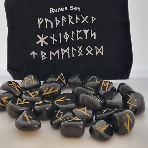 Black agate rune stones with golden symbols scattered in front of a black pouch featuring runic designs, showcasing their smooth surfaces and intricate markings.