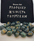 Bloodstone rune stones with golden symbols scattered in front of a black pouch featuring runic designs, showcasing their smooth surfaces and deep green with red speckled coloring.