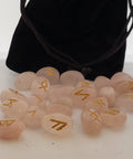 Rose quartz rune stones with golden symbols scattered in front of a black pouch featuring runic designs, showcasing their smooth, polished surfaces and soft pink color.